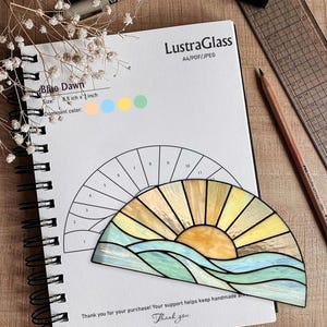 May include: A stained glass sun and ocean design, with blue, yellow, and orange hues, rests on a spiral-bound notebook. The notebook has the text "Blue Dawn" and "LustraGlass" printed on it. The size is 8.5 inches x 7 inches. The image includes pens and a ruler.