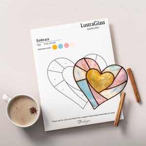 May include: A heart-shaped stained glass art piece with a yellow centre and pastel blue, pink, and white sections, next to a printed template. The template is labelled "Embrace" and is 20 cm by 20 cm. A cup of coffee and cinnamon sticks are also present.