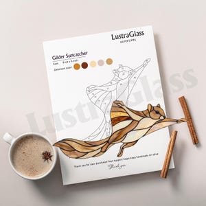 May include: A Glider Suncatcher design on a white paper, with the text "LustraGlass". The design features a flying squirrel with brown and cream colors. The size is 8 inches x 5 inches. Cinnamon sticks and a cup of coffee are also present.