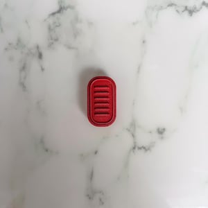 Puede incluir: Un pequeño cortador de galletas rojo rectangular con bordes redondeados y una serie de líneas paralelas. El cortador está hecho de un material sólido y está sobre un fondo de mármol blanco y gris.