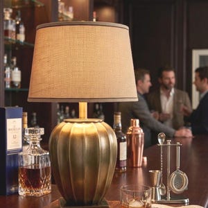 Puede incluir: Una lámpara de mesa de latón con una pantalla de tela beige, colocada en una barra con un vaso de whisky y herramientas de bar. La lámpara tiene una base redondeada y acanalada. Otros artículos incluyen una jarra, una botella de whisky y una caja de cerillas.