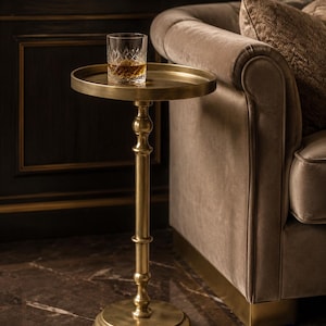 Puede incluir: Una mesa auxiliar redonda y alta fabricada en metal de latón cepillado con una base de pedestal decorativa y esbelta y una superficie circular tipo bandeja. Un vaso de cristal tallado con un líquido color ámbar descansa sobre la mesa.