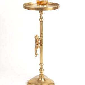 May include: A gold-coloured side table with a round tray top. A small glass with a drink and a stirring spoon sits on the tray. A decorative monkey climbs the table's central pole. The base is a four-leaf clover shape.