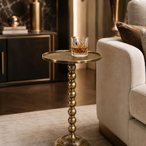 Op de afbeelding: Een kleine, antieke messing bijzettafel met een rond blad en een stam met kralen. Een geslepen kristallen glas met amberkleurige vloeistof staat op de tafel. De tafel staat naast een beige bank.