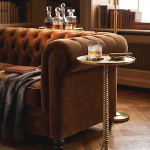 May include: A brown velvet sofa with a side table holding a glass of amber liquid and a cigar. The side table has a gold-coloured, twisted stem. A brass lamp and bottles of liquor are visible in the background.