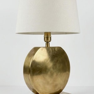 Peut inclure: Une lampe de table dorée avec un abat-jour crème. Le pied de la lampe a une forme sphérique avec des facettes géométriques, reposant sur une base rectangulaire. L'abat-jour est un cylindre simple et conique.