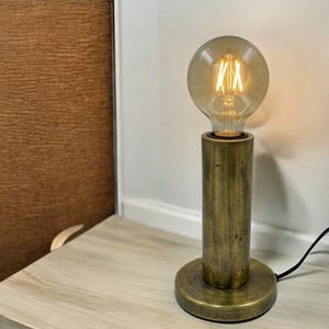 Op de afbeelding: Een vintage tafellamp met een grote, ronde Edison-stijl gloeilamp. De lamp heeft een cilindrische basis en een cirkelvormige basis, beide met een messing afwerking. De lamp geeft warm licht. Een zwart netsnoer is aangesloten. De lamp is een decoratieve verlichtingsarmatuur.