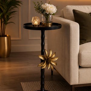 May include: A black metal accent table with a round top and a slender, textured stem. The base is circular, and the stem features a decorative gold-colored metal floral element. A glass vase with white flowers and a small glass candle holder sit on top.