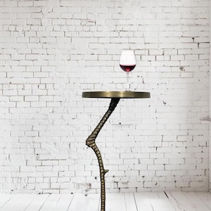 May include: A unique side table with a bronze finish, featuring a bird's foot base and a curved leg. The round tabletop holds a glass of red wine. The table is set against a white brick wall and a white wooden floor.