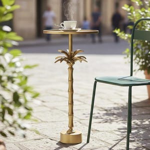Puede incluir: Una mesa auxiliar dorada con un diseño de palmera que sostiene una mesa redonda. Una taza y un platillo blancos están sobre la mesa. La mesa está al aire libre, junto a una silla verde y plantas en macetas.