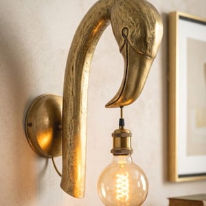 May include: A brass-colored wall sconce shaped like a flamingo's head and neck. The light fixture holds a round, Edison-style lightbulb. The sconce is mounted on a wall, with a framed picture visible in the background.