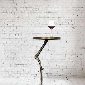 以下が含まれることがあります： 鳥の足のようなデザインの、ユニークなブロンズ色のサイドテーブル。テーブルは丸い天板で、赤ワインのグラスが置かれています。白いレンガの壁と白い木の床を背景にテーブルが置かれています。