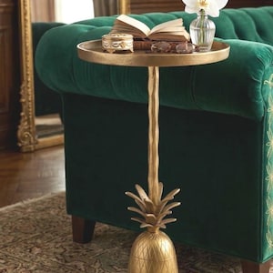 May include: A gold-colored side table with a pineapple base and a round top. The table holds a small decorative box, an open book, and a vase with a white orchid. The table is next to a green velvet sofa.