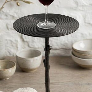 Pode incluir: Um suporte de taça de vinho de metal preto com um tampo redondo e uma haste em forma de galho de árvore. Uma taça de vinho cheia de vinho tinto está no topo. O suporte está em uma superfície de madeira.