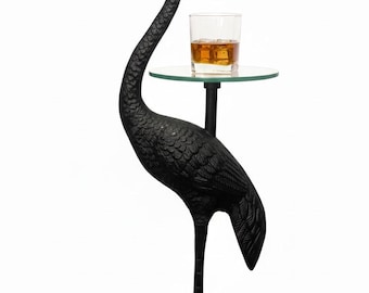 May include: A black metal crane sculpture with a small glass shelf holding a glass of amber liquid. The crane has a long, curved neck and stands on a round base. The shelf is round and clear.