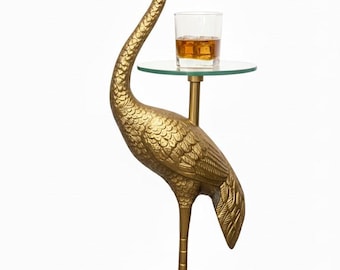 May include: A gold-coloured crane sculpture with a glass shelf holding a glass of amber liquid. The crane has detailed feather and scale textures. The sculpture stands on a round base and has a long, curved neck.