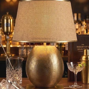 May include: A gold table lamp with a textured, egg-shaped base and a tan lampshade. The lamp sits on a rectangular base. A cocktail glass, ice, and bar tools are in the foreground. The background shows a bar setting.