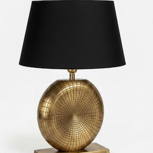 May include: A decorative table lamp with a black lampshade and a gold-colored base. The lamp's base is spherical with a textured, grid-like pattern. The lamp sits on a rectangular base, adding a touch of elegance to any room.