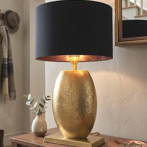 May include: A gold-coloured table lamp with a textured, oval base and a black lampshade. The lampshade has a gold interior. The lamp sits on a rectangular base. A small vase with greenery is next to the lamp.