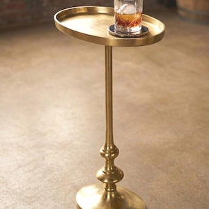 May include: A gold-coloured, oval-shaped side table with a tall, slender stem and a round base. A glass of amber liquid with ice cubes sits on the table's surface. The table has a decorative, ornate design.