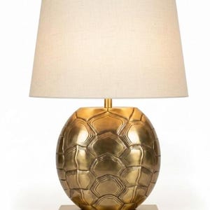 May include: A gold-colored table lamp with a cream-colored lampshade. The lamp base is spherical with a textured, geometric pattern. The base sits on a square, gold-colored platform. The lampshade is a classic, tapered shape.