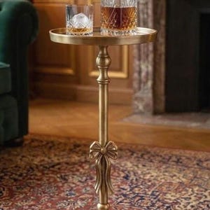 Puede incluir: Una mesa auxiliar dorada con una tapa redonda, que contiene una jarra de cristal y un vaso de whisky con hielo. La mesa tiene un diseño de lazo decorativo y se apoya en una alfombra estampada.