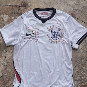 Può includere: Maglia da calcio bianca con rifiniture blu navy e scollo a V. Presenta lo stemma dell'Inghilterra, ricami floreali e il logo Nike. Il testo "HAPPY AND GLORIOUS" è stampato sul colletto.