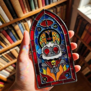 Könnte beinhalten: Ein Buntglasfenster mit roter, blauer und gelber Farbgebung. Das Panel zeigt einen Cartoon-Lamm-Charakter mit roten Augen, Flammen und einem Totenkopf-Anhänger. Das Panel wird in einer Hand gehalten.