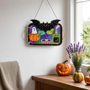 Könnte beinhalten: Ein dekoratives Tablett zum Thema Halloween mit einem Design aus schwarzer Fledermaus, Geist und Kürbis. Das Tablett hat einen goldfarbenen Perlenbesatz und wird an einer Kette aufgehängt. Das Bild zeigt auch geschnitzte Kürbisse und eine Vase mit Blumen.