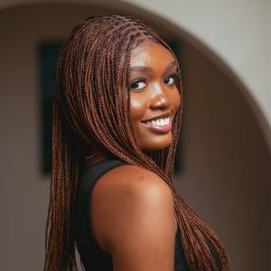 May include: A woman with long, brown braided hair smiles at the camera. The braids are finely woven and cascade down the shoulders. She wears a black top. The background is blurred, with neutral tones.