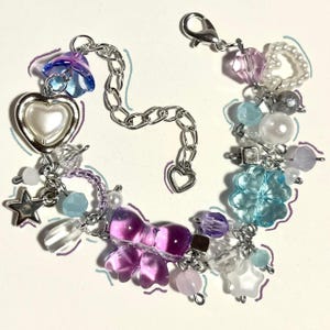 May include: A silver charm bracelet adorned with a selection of colourful beads and charms. The bracelet includes a heart-shaped charm, a star, a bow, and various translucent and opaque beads in shades of purple, blue, and white.