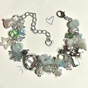 May include: A silver charm bracelet with a mix of beads and charms. It includes light blue, green, and clear beads, pearl accents, star charms, and a heart-shaped charm. The bracelet is set against a white backdrop.