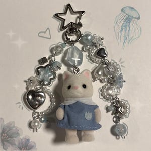 May include: A keychain featuring a white flocked cat figurine wearing a blue dress, suspended from a silver star-shaped clasp. The cat is flanked by two beaded strands containing clear, blue, and silver-toned charms, including hearts, stars, and pearls.