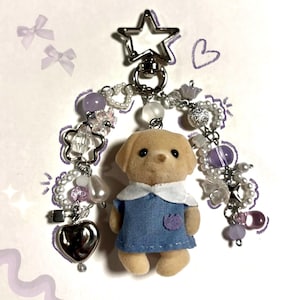 May include: A keychain featuring a flocked tan dog figurine wearing a blue dress with a white collar, suspended from a silver star-shaped clasp. The charm is decorated with two strands of beads, including purple spheres, white pearls, and silver hearts.
