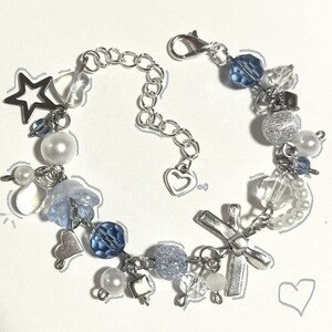 Puede incluir: Una pulsera delicada con una mezcla de cuentas azules, transparentes y plateadas. Incluye un dije de estrella, dijes en forma de corazón y un colgante de cruz. El diseño incorpora perlas y varias formas de cuentas, conectadas por una cadena plateada.