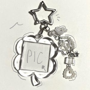May include: Clear acrylic keychain with a clover-shaped frame and the word "PIC" written inside. The keychain features a star-shaped clasp and decorative charms, including a flower, pearls, and a heart-shaped cluster of beads. The design is delicate and feminine.