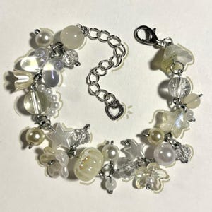 Könnte beinhalten: Ein filigranes Armband mit einer Mischung aus weißen und irisierenden Perlen, darunter Perlen, Sterne und Blumenformen. Das Armband hat eine silberne Kette mit Herzanhänger und einem Karabinerverschluss.