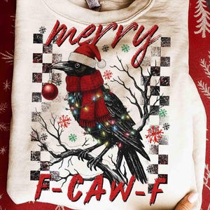 Könnte beinhalten: Cremefarbenes Sweatshirt mit Weihnachtsgrafik. Das Design zeigt eine schwarze Krähe mit Weihnachtsmütze, rotem Schal und Lichterkette, die eine rote Kugel hält. Die Wörter "merry" und "F-CAW-F" sind ebenfalls abgebildet.