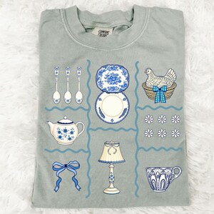 Pode incluir: Camiseta verde s&aacute;lvia com um design de grade com ilustra&ccedil;&otilde;es de uma chaleira, pratos, colheres, uma l&acirc;mpada, uma x&iacute;cara de ch&aacute;, um la&ccedil;o e uma galinha em uma cesta. As ilustra&ccedil;&otilde;es s&atilde;o em azul e branco.