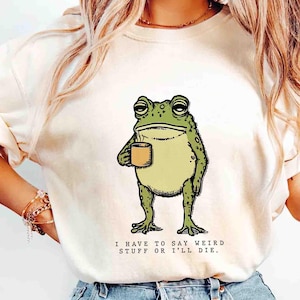 May include: Cream-colored t-shirt featuring a cartoon frog holding a coffee cup. The frog is green with a light green belly. Text below the frog reads, "I HAVE TO SAY WEIRD STUFF OR I'LL DIE."