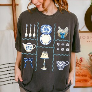 Pode incluir: T-shirt cinzenta escura com um design de grelha com v&aacute;rias ilustra&ccedil;&otilde;es. O design inclui ch&aacute;venas, pratos, colheres, uma chaleira, uma l&acirc;mpada, uma galinha numa cesta e la&ccedil;os, todos em azul e branco. A t-shirt tem um aspeto vintage.