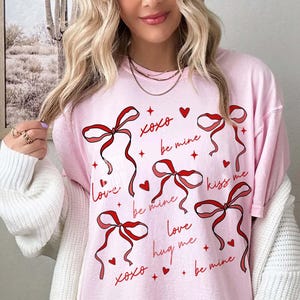May include: Pink t-shirt with red ribbon graphics and romantic phrases like "Love," "Be Mine," and "Kiss Me." The design includes hearts and stars, creating a Valentine's Day or romantic theme.