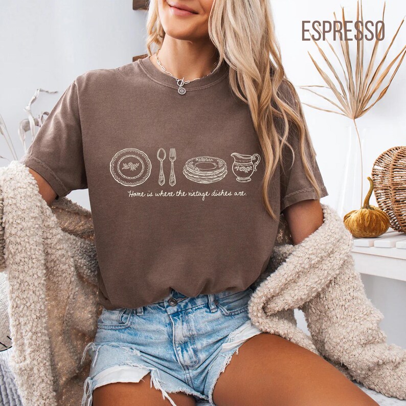 May include: Espresso-colored t-shirt with a white graphic of dishes, silverware, and a creamer. The text "Home is where the vintage dishes are" is below the graphic. The shirt is short-sleeved and worn with denim shorts.
