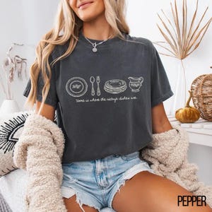 May include: Dark gray t-shirt with a white graphic of vintage dishes and the text "Home is where the vintage dishes are." The shirt is a comfortable, casual style, perfect for everyday wear. The shirt is made of a soft material.