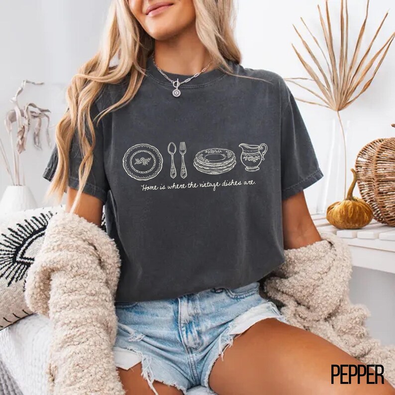 May include: Dark gray t-shirt with a white graphic of vintage dishes and the text "Home is where the vintage dishes are." The shirt is a comfortable, casual style, perfect for everyday wear. The shirt is made of a soft material.