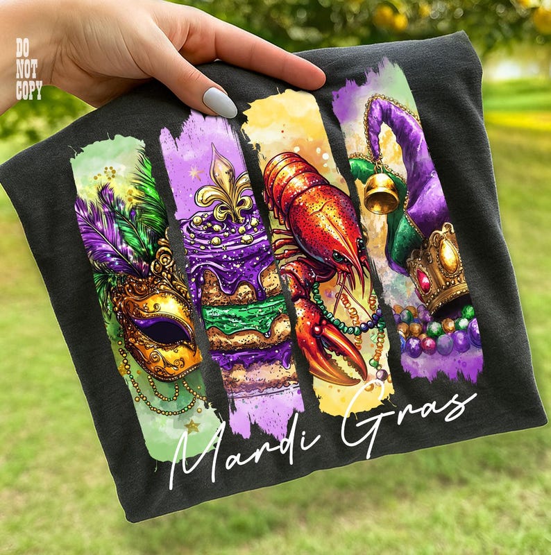 May include: Dark gray t-shirt featuring a colorful Mardi Gras design. The design includes a mask, cake, crawfish, and jester hat with the words "Mardi Gras" in white script. The shirt is held up by a hand.