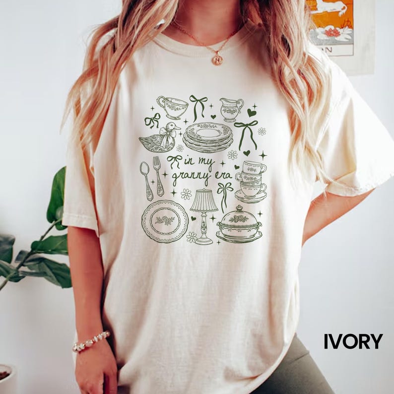 May include: Ivory t-shirt with a green illustration of kitchen items like plates and cups, and a duck, with the text "in my granny era." The shirt has a relaxed fit. The word "IVORY" is printed in bold, capital letters.
