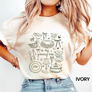May include: Ivory-coloured t-shirt with a green line drawing of vintage kitchenware, including plates, teacups, a duck, and the text "in my granny era." The shirt has a crew neck and short sleeves. The word "IVORY" is printed in black.