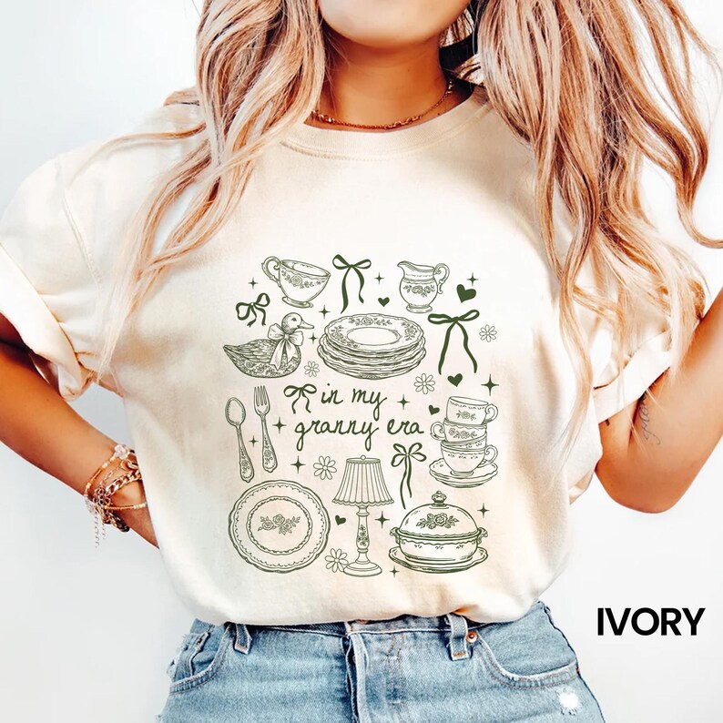 May include: Ivory-coloured t-shirt with a green line drawing of vintage kitchenware, including plates, teacups, a duck, and the text "in my granny era." The shirt has a crew neck and short sleeves. The word "IVORY" is printed in black.