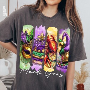 May include: Dark gray t-shirt featuring a colorful Mardi Gras design. The design includes a mask, cake, crawfish, and jester hat, with the words "Mardi Gras" in white script. The shirt is a comfortable, casual style.